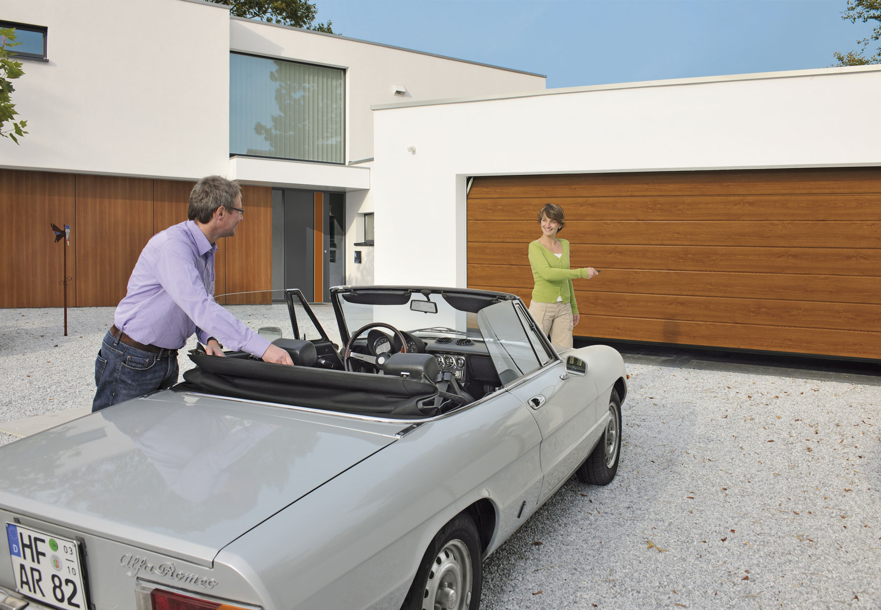 Ein Cabrio steht in einer Einfahrt und eine Frau betätigt den Sensor damit sich das Holzgaragentor öffnet.